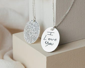 May include: Two silver necklaces with oval pendants. One pendant features a detailed fingerprint texture, while the other displays the engraved handwritten text "I Love You". Both pendants hang from delicate silver chains on light-colored blocks.
