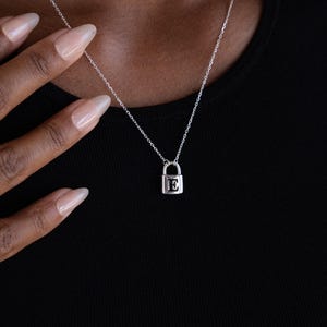 May include: A silver-colored padlock pendant necklace with a delicate chain. The pendant features the letter "E" inside the lock. The necklace is worn against a black top.