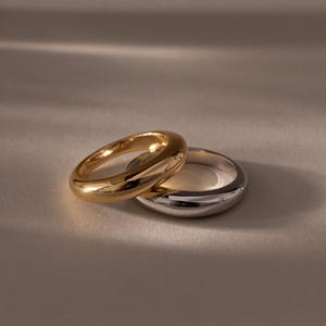 Può includere: Due anelli, uno dorato e uno argentato, sono presentati insieme. L'anello dorato ha una finitura lucida e brillante. L'anello argentato ha un design e una finitura simili. Gli anelli sono su uno sfondo neutro, con ombre morbide.