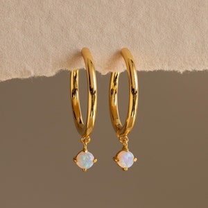 Puede incluir: Un par de pendientes de aro de oro con una pequeña opala blanca colgando de cada aro.