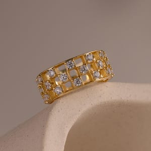 May include: A gold-colored ring with a geometric design. The ring features a series of square cutouts and is adorned with small, clear gemstones. The ring is displayed on a neutral-toned surface, showcasing its intricate details and craftsmanship.