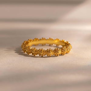 May include: A gold-toned ring featuring a continuous band of small, repeating flower-shaped motifs. The metal surface has a polished finish, and the design consists of a series of interconnected floral elements that form the circular band of the ring.