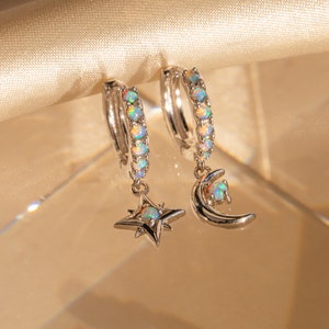 May include: A pair of silver hoop earrings with a dangling star and crescent moon charm. The hoops are adorned with iridescent opal stones.