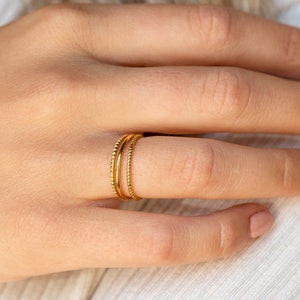 A close up of our Dainty Stacking Ring Set in 18K Gold finish placed on on models ring finger - featuring a set of 3 rings: 1 Thin Band, 1 Twist Ring and 1 Notched Ring all about 1mm thick.
