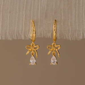May include: A pair of gold-colored hoop earrings with bow-shaped charms and teardrop-shaped clear gemstones. The earrings are displayed against a neutral-toned background, highlighting their delicate design and sparkle.