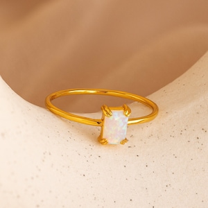 May include: A gold-colored ring with a rectangular, iridescent opal stone. The ring's band is thin and delicate, and the opal is set in a four-prong setting. The ring is displayed on a neutral-toned surface.