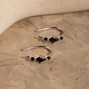 Puede incluir: Un par de pendientes de aro de plata con tres piedras preciosas negras dispuestas en forma de triángulo.