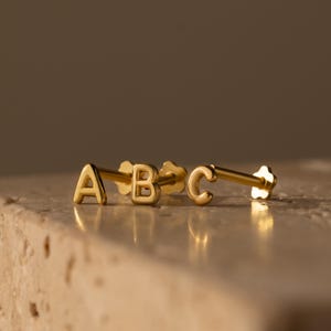 Może przedstawiać: Złote kolczyki z literami "ABC". Kolczyki są prezentowane na jasnej, teksturowanej powierzchni. Litery są w prostej, bezszeryfowej czcionce.