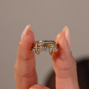 May include: A stack of three rings, held between fingers, featuring a mix of silver and gold tones. The top and bottom rings have a silver band with small, clear stones. The middle ring is gold with a twisted rope design. Two gold bars connect the rings.