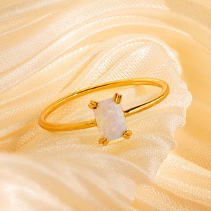 May include: A gold-colored ring with a rectangular, iridescent opal stone. The ring is set on a soft, cream-colored fabric, highlighting the ring's delicate design. The opal stone is the focal point, showcasing its subtle color variations.