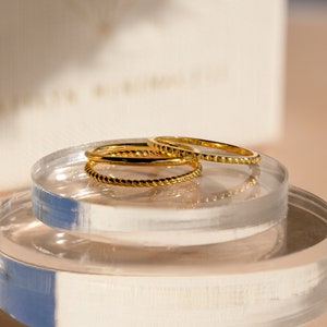 A close up of our Dainty Stacking Ring Set in 18K Gold finish placed on a clear decorative dish with our Caitlynminimalist gift box in the background - featuring a set of 3 rings: 1 Thin Band, 1 Twist Ring and 1 Notched Ring all about 1mm thick.