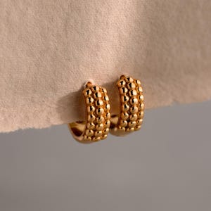 May include: A pair of gold-colored hoop earrings. The earrings have a textured surface with a series of small, round, raised bumps. The earrings are displayed against a neutral-toned background.