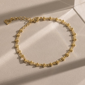 May include: A delicate gold bracelet with a double-strand design. The bracelet features small, evenly spaced beads and a heart-shaped charm near the clasp. The jewelry is displayed on a light-colored surface.