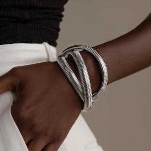 May include: A silver-colored bracelet with a coiled design is worn on a person's wrist. The bracelet wraps around the wrist multiple times, creating a layered effect. The person is wearing a black top and white pants.