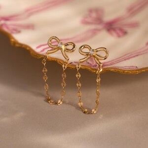 Bow Chain Stud Earrings in gold featuring delicate bow studs with a central stone and draped chain loops, displayed on a decorative scalloped dish with pink ribbon design.