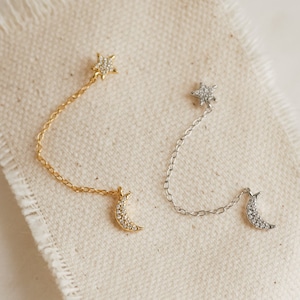 A gold earring and a silver earring lay flat on a beige piece of canvas. Both earrings have the same design; a pave star stud at the top and a pave moon stud at the bottom, connected by a dainty chain.