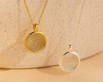 May include: Two necklaces with round pendants, one in gold and one in silver. Each pendant features a detailed, engraved fingerprint design on the front surface. The pendants hang from delicate matching metal chains with a simple link structure.