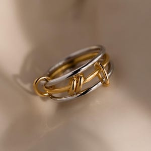 Op de afbeelding: Een tweekleurige ring met een goud- en zilverontwerp. De ring heeft een uniek knoopachtig detail in het midden, met een gouden band verweven met zilveren banden. De ring is een sieraad.