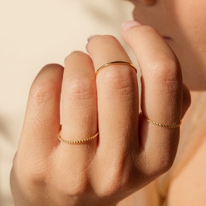Puede incluir: Una mano con tres anillos de oro. Los anillos son finos y delicados, uno de ellos con un diseño trenzado.