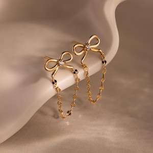 Puede incluir: Un par de pendientes dorados con forma de lazo y un delicado diseño de cadena. Cada pendiente presenta un lazo en la parte superior con una pequeña gema y una cadena de pequeños abalorios redondos. Los pendientes se exhiben sobre una superficie blanca.
