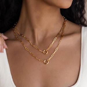 May include: Two gold-colored chain necklaces. The necklaces are layered, with a shorter chain above a longer one. Each chain features a series of rectangular links and a circular clasp. The jewelry is worn over a white tank top.