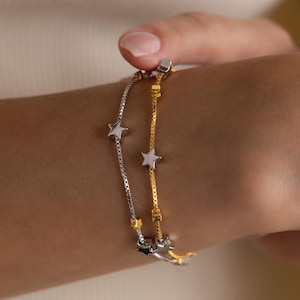 Puede incluir: Dos pulseras delicadas, una plateada y otra dorada, cada una adornada con abalorios en forma de estrella. Las pulseras se muestran sobre un fondo neutro, mostrando sus finos detalles y su elegante diseño. Un accesorio elegante para el uso diario.
