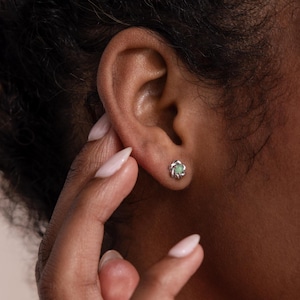 May include: A close-up of a silver stud earring with a flower design. The earring has a light green center and is set against the ear of a person with dark skin. The person's hand is near the ear.