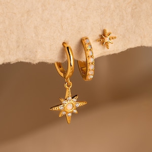 May include: A set of three gold earrings featuring a starburst design with opal accents. The earrings are on a beige background.