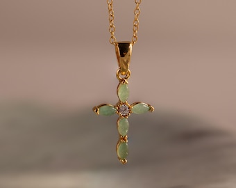 May include: A gold-colored necklace with a cross pendant. The cross is adorned with light green oval-shaped stones and a small, sparkling center stone. The necklace hangs against a neutral background.
