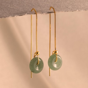 May include: A pair of gold-coloured threader earrings. Each earring has a small, round, pale green stone with a hole in the middle. The earrings hang from a thin gold chain.