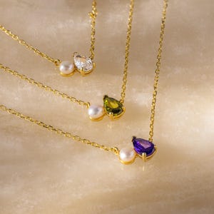 May include: Close-up of three gold necklaces with different gemstone pendants. Each necklace features a small pearl paired with a pear-shaped gemstone in clear, green, and purple. The necklaces are displayed on a light-colored, textured surface.