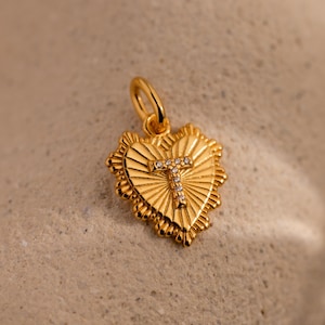 May include: A gold-colored heart-shaped pendant with a sunburst design and a letter "T" encrusted with small, clear gemstones. The pendant has a small loop at the top for a chain.