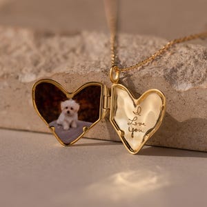 May include: Gold heart-shaped locket necklace, open to reveal a small photo of a white dog. The locket is engraved with "I Love You" on the front. The necklace is displayed on a textured, neutral-colored surface.