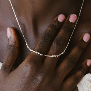 Könnte beinhalten: Eine silberne Halskette mit einer zarten Kette und einem geschwungenen Balken-Design mit kleinen Perlen. Die Halskette wird von einer Person mit dunkler Haut und rosafarbenem Nagellack getragen.