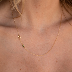 A close up of our Sideways Initial Birthstone Necklace in 18K Gold finish on our models neck hanging just below her collarbone - featuring a small 7mm letter S charm and a 3mm green emerald stone hanging sideways on a dainty cable chain.