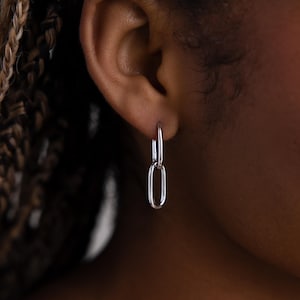 Op de afbeelding: Een zilveren oorbel met een schakelkettingontwerp. De oorbel is een hoepel met een hangende, langwerpige ovale vorm. De oorbel wordt gedragen op het oor van een persoon, met hun huid en haar zichtbaar op de achtergrond.