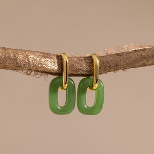 May include: A pair of gold-tone hoop earrings featuring rectangular, translucent green resin charms. The earrings are suspended from a textured, metallic brown branch-like element, showcasing a polished finish on the metal hoops.