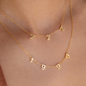 以下が含まれることがあります： 数字が付いた2つの繊細なゴールドネックレス。上のネックレスには数字の2、2、1が、下のネックレスには数字の7、9、9、6が付いています。ネックレスは首の周りに着用されています。