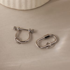 May include: Two silver-colored metal jewelry clasps. One is a U-shaped shackle with a screw closure, and the other is an oval-shaped carabiner with a screw closure. Both clasps have a shiny, reflective surface.