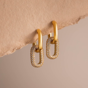 May include: A pair of gold-toned hoop earrings featuring a dangling rectangular link charm. Each link is encrusted with small, clear, sparkling stones along its entire perimeter, creating a bright, reflective surface against the smooth gold hoops.