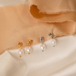 May include: Close-up of four earrings with a pearl drop. Two earrings have gold-colored heart-shaped tops, and two have silver-colored tops. The earrings are displayed on a white and tan fabric background.