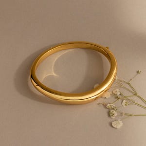 May include: A polished gold-tone metal bangle bracelet with an irregular, organic, and rounded shape. The bracelet features a smooth, reflective surface and a hinged clasp mechanism, displayed alongside a small sprig of dried white baby's breath flowers.