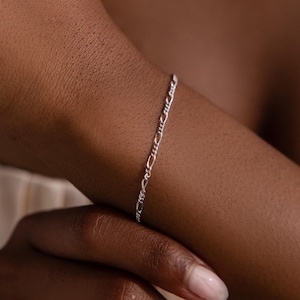 May include: A delicate silver Figaro chain bracelet. The bracelet features a repeating pattern of three small links followed by one elongated link. The bracelet is worn on a wrist.