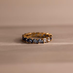 May include: A gold ring with a band of alternating blue and clear marquise-cut gemstones. The ring is set against a neutral background, highlighting the sparkle of the stones. The ring's design is elegant and suitable for various occasions.