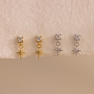 May include: Gold and silver-toned stud earrings with a dangling starburst design. Each earring features a small, clear gemstone at the top. The starburst pendants hang below the gemstone studs. The earrings are displayed on a neutral background.