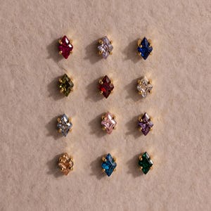 May include: A collection of twelve gemstone stud earrings in various colors, including red, blue, green, and pink. Each earring features a diamond-shaped gemstone set in a gold-colored metal setting. The earrings are arranged in a grid pattern on a neutral-toned surface.