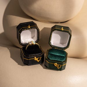 May include: Two open jewelry boxes, one black and one green, sit on a neutral surface. The black box contains a gold ring with a gemstone. The green box has a green velvet interior. Both boxes have gold accents and clasps.