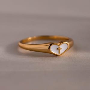 Puede incluir: Anillo dorado con forma de corazón. El corazón es blanco con una cruz dorada en el centro. El anillo tiene un acabado liso y pulido. El fondo es de color neutro.
