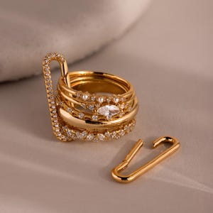 May include: A collection of gold rings, some adorned with small, clear gemstones, and a gold safety pin. The rings are stacked together, and the safety pin is open, creating a unique jewelry set.