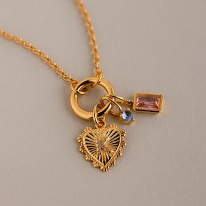 Könnte beinhalten: Goldfarbene Halskette mit herzförmigem Anhänger, rechteckigem rosa Edelstein und kleinem blauen Edelstein. Der Herzanhänger hat ein strukturiertes Design mit einem kleinen Kristall. Auf neutralem Hintergrund.